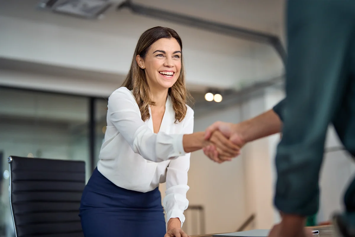 Happy mid aged business woman manager handshaking greeting client in office. Smiling female executive making successful deal with partner shaking hand at work standing at meeting table.